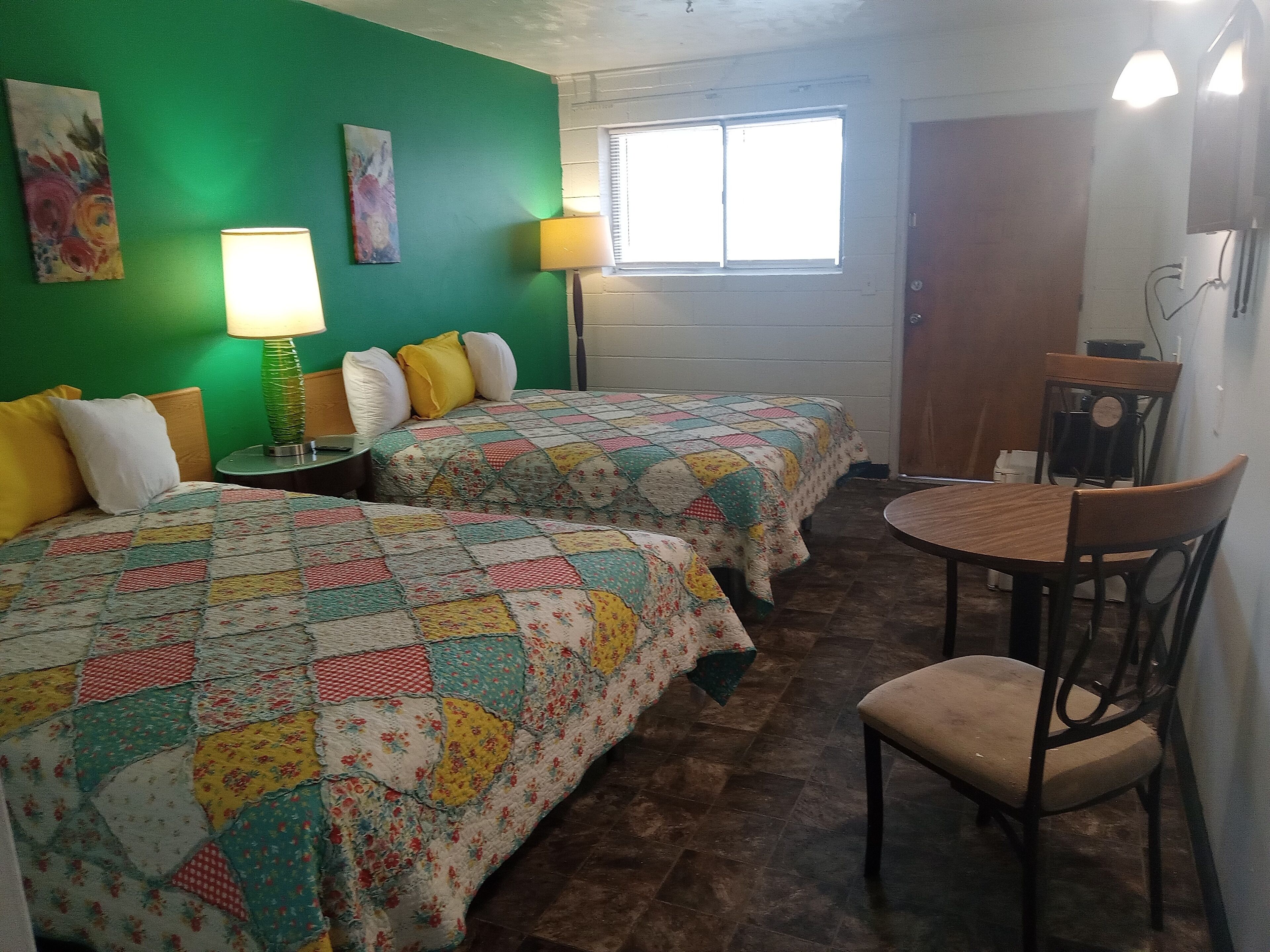 Una habitación con dos camas, una mesa y sillas. Hay dos lámparas en las mesitas de noche.