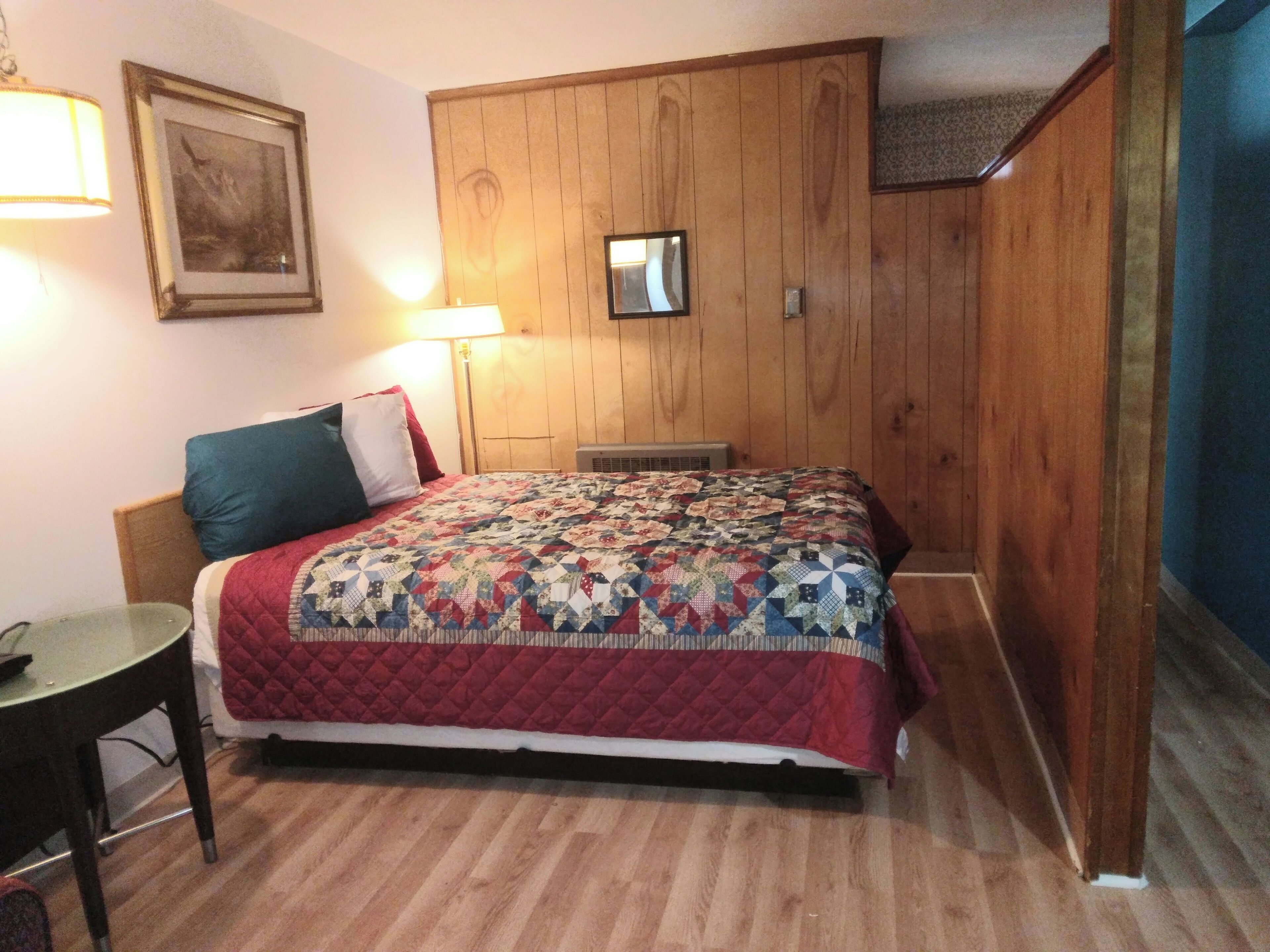 A bedroom with a wooden wardrobe, a bed with a quilt, a bedside table, and a lamp.