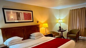 A hotel room with a large bed, a nightstand, a desk with a lamp, a chair, and a framed picture on the wall. - Ceilidh Country Lodge (Baddeck)