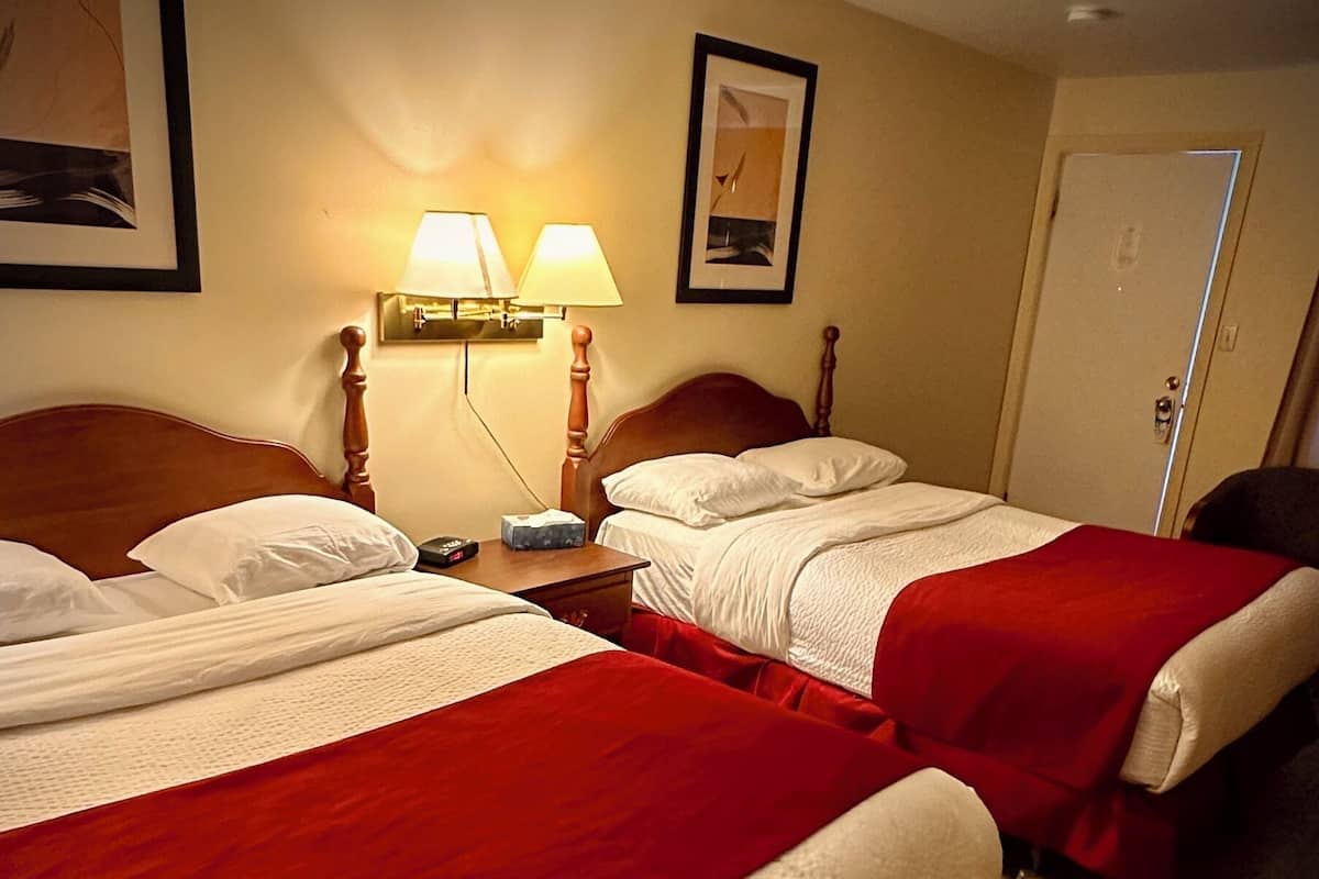 A hotel room with two beds, each with a red blanket and white bedding. There are two wall-mounted lamps, a framed picture on the wall, and a door to the right.