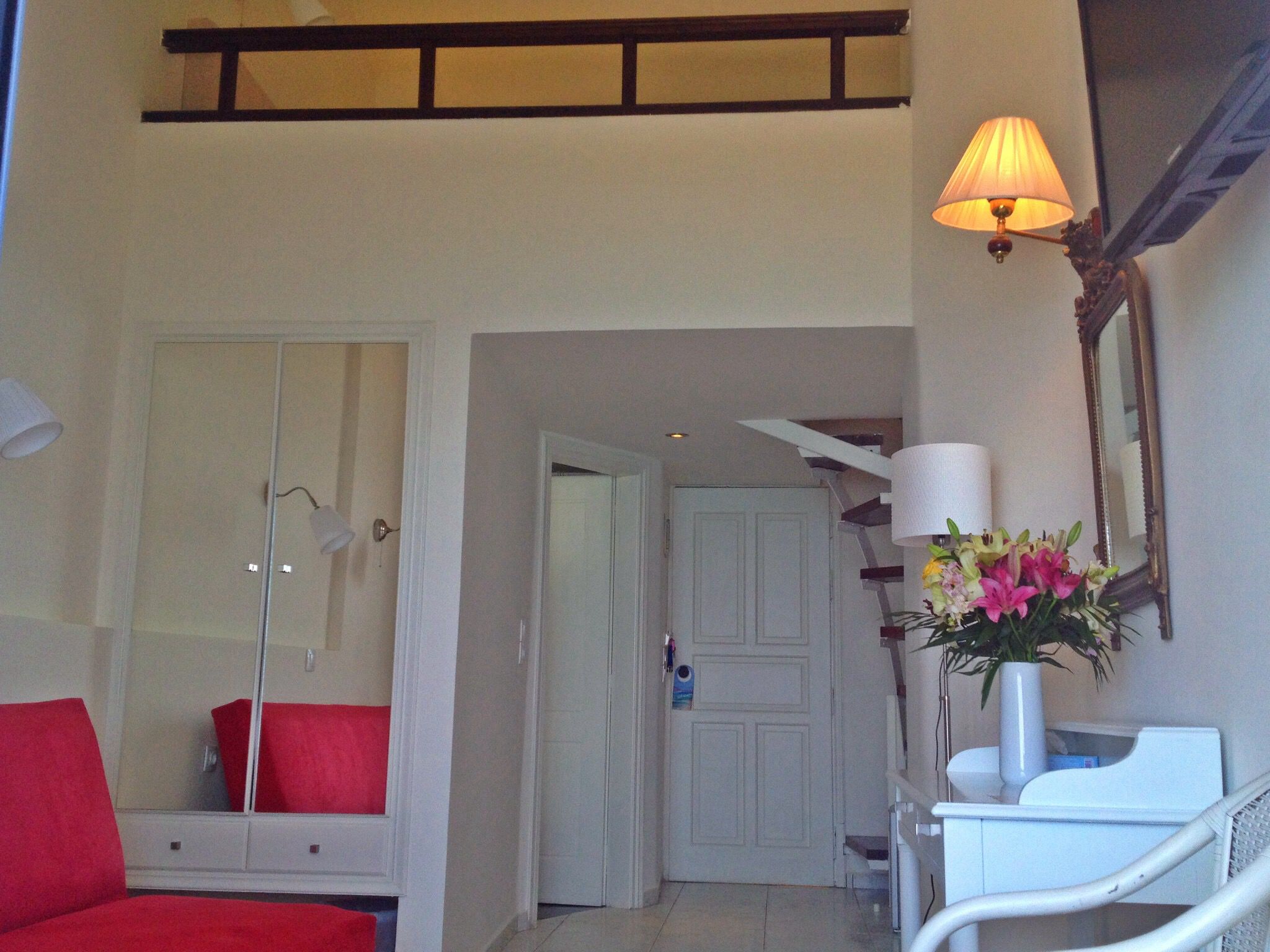 Una habitación de hotel con un espejo grande, una silla roja, una mesita de noche blanca con un jarrón de flores y una escalera que lleva a otra estancia.