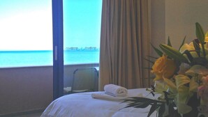 Une chambre d’hôtel avec un lit, une chaise, une fenêtre donnant sur la mer et un vase de fleurs.