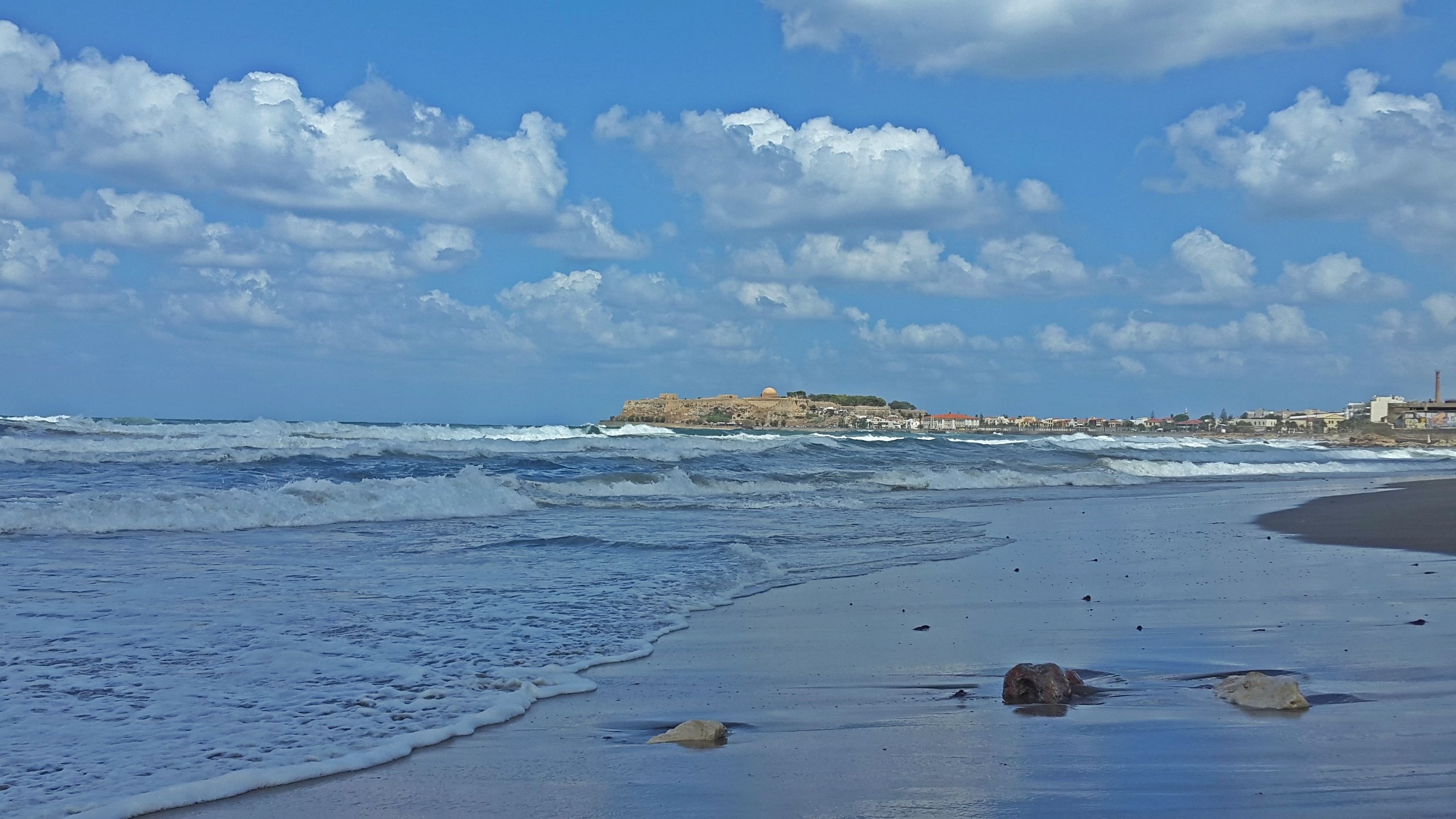 Una spiaggia con onde, una riva sabbiosa e una struttura rocciosa in lontananza con degli edifici.