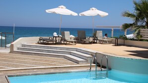 Un espace aménagé au bord de la piscine, avec des chaises longues et des parasols, une vue dégagée sur l’océan et une piscine dotée d’un escalier en métal.