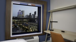A hotel room with a large window offering a city view, a desk with a lamp, and a bed with a patterned headboard.