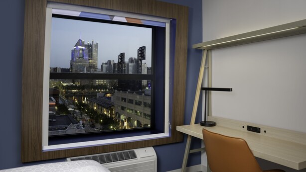 A hotel room with a large window offering a city view, a desk with a lamp, and a bed with a patterned headboard. - Holiday Inn Express & Suites Pittsburgh North Shore by IHG (Pittsburgh)
