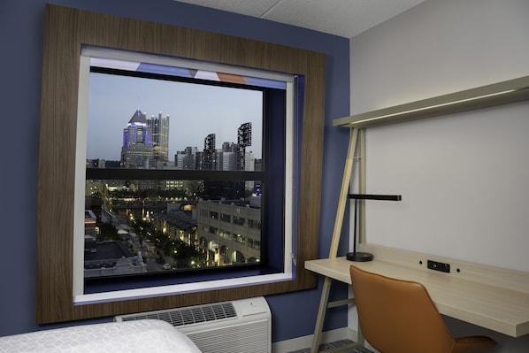 Habitación de hotel con un ventanal grande que ofrece vistas a la ciudad, un escritorio con lámpara y una cama con cabecero estampado.