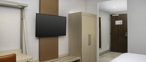 Habitación de hotel con cama, televisor montado en la pared, armario de madera, banco y puerta.