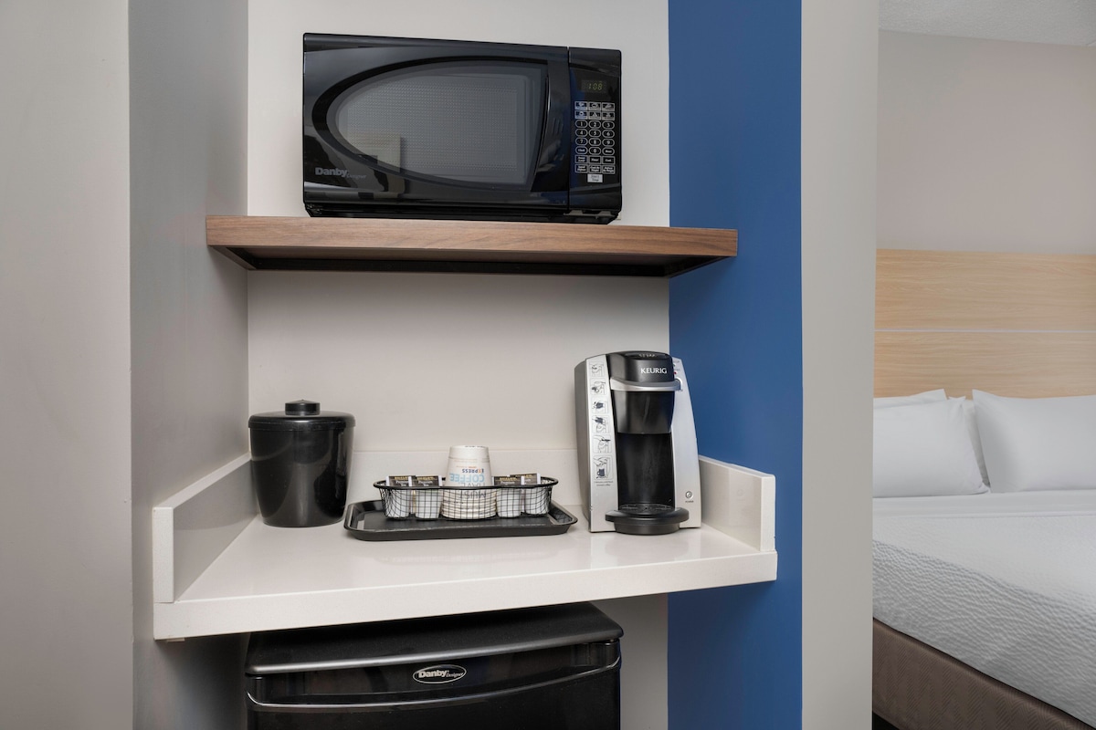 A hotel room with a microwave on a shelf, a coffee maker, and a trash bin.