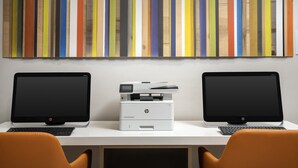 Two HP desktop computers on a white desk with an HP printer between them, and a colorful striped wall art above.