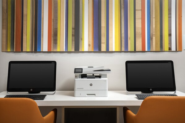 Dos computadoras de escritorio HP sobre un escritorio blanco, con una impresora HP en medio, y un cuadro de pared a rayas coloridas encima.