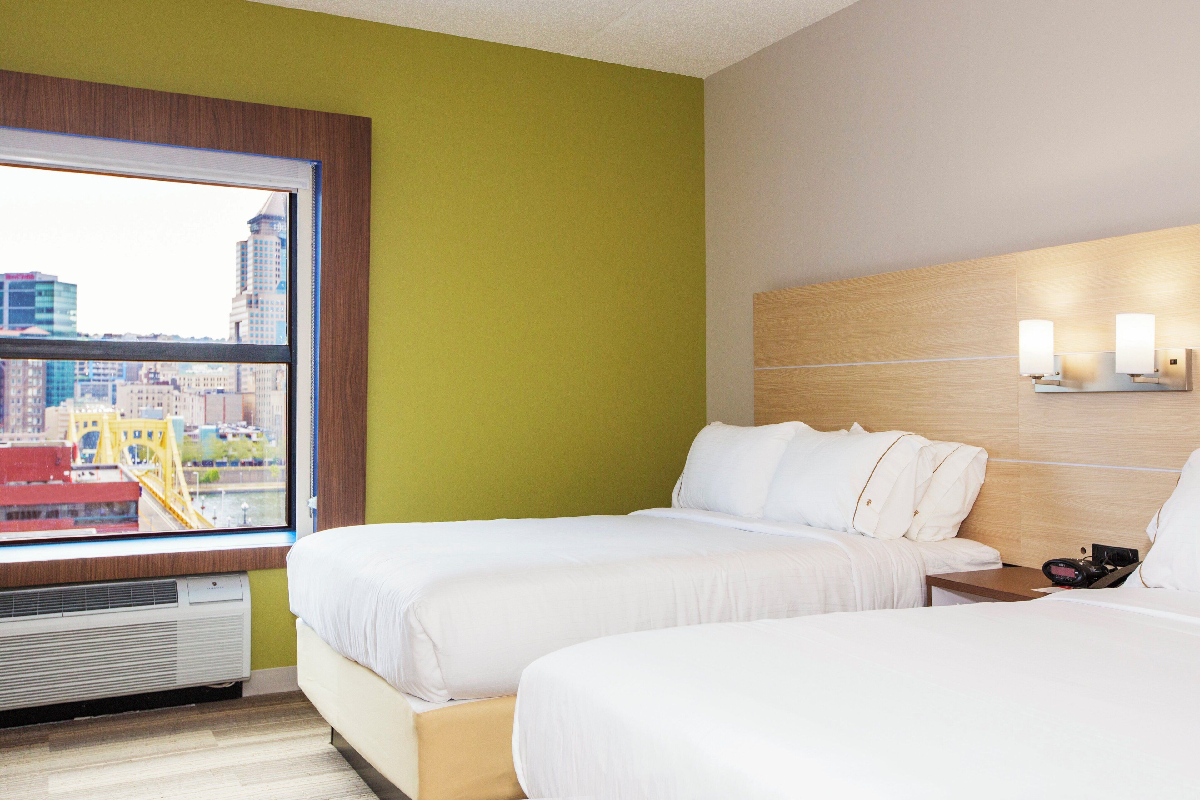 A hotel room with two beds, a window with a view of a cityscape, and a wall-mounted light fixture.