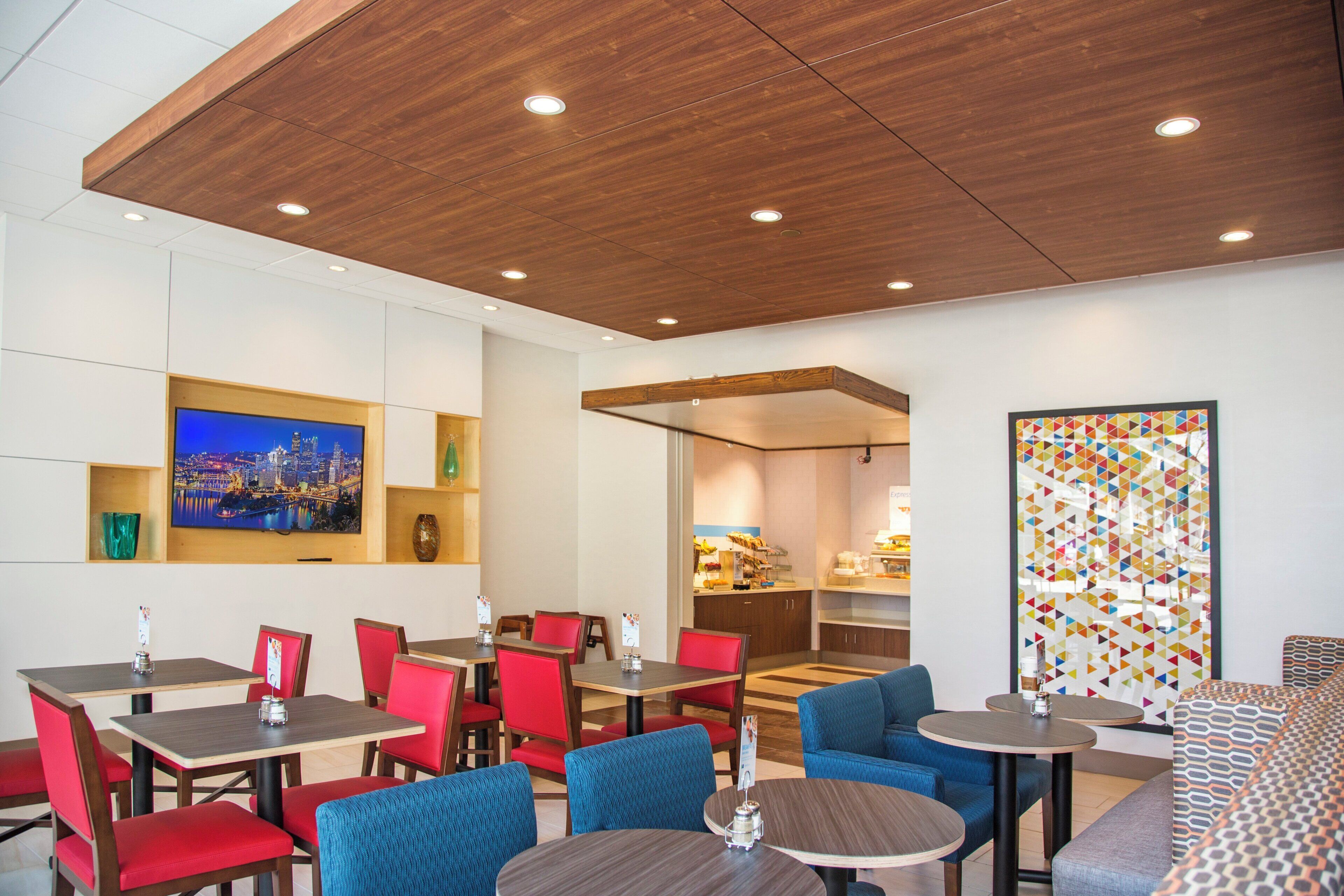 Um espaço de refeições de hotel com várias mesas e cadeiras, uma televisão exibindo uma vista urbana e um design de parede geométrico e colorido.