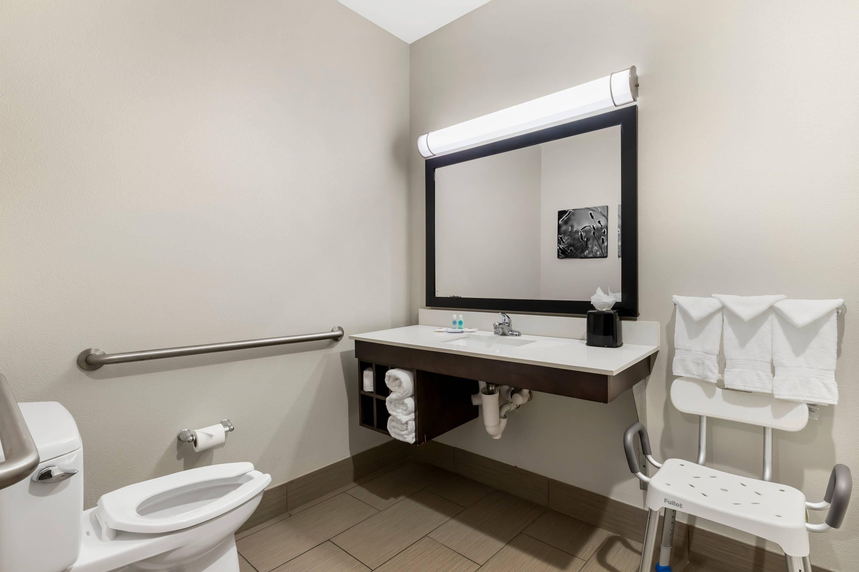 A modern bathroom with a toilet, sink, mirror, towel rack, and a wheelchair-accessible chair.