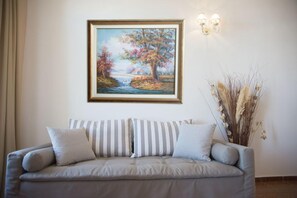 A neutral-toned sofa with striped cushions, a framed landscape painting above it, and a vase with dried plants to the right. - Porto Enetiko Suites (Rethymno)