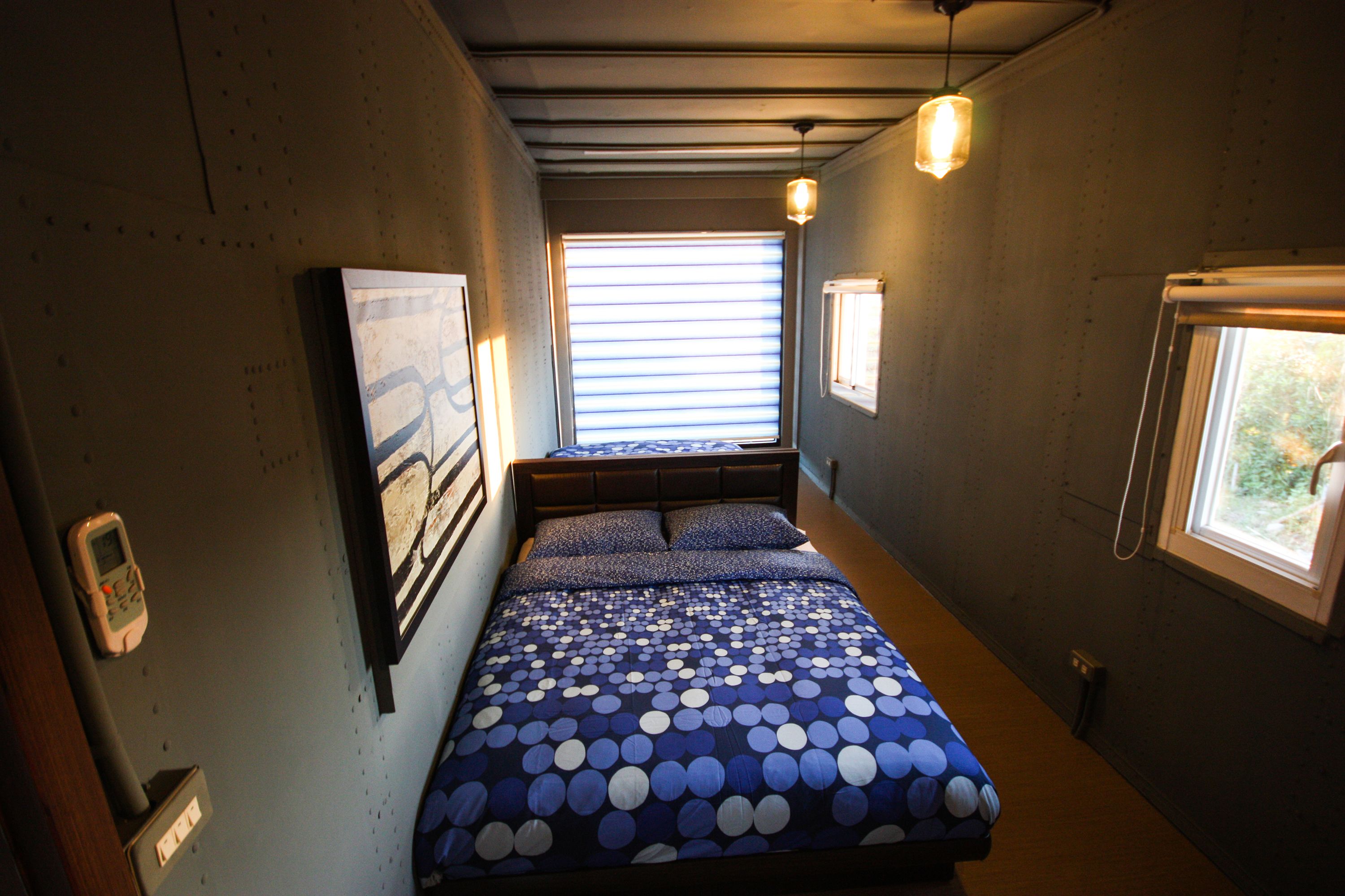 Una habitación estrecha con una cama, un ventanal y una lámpara fijada a la pared.