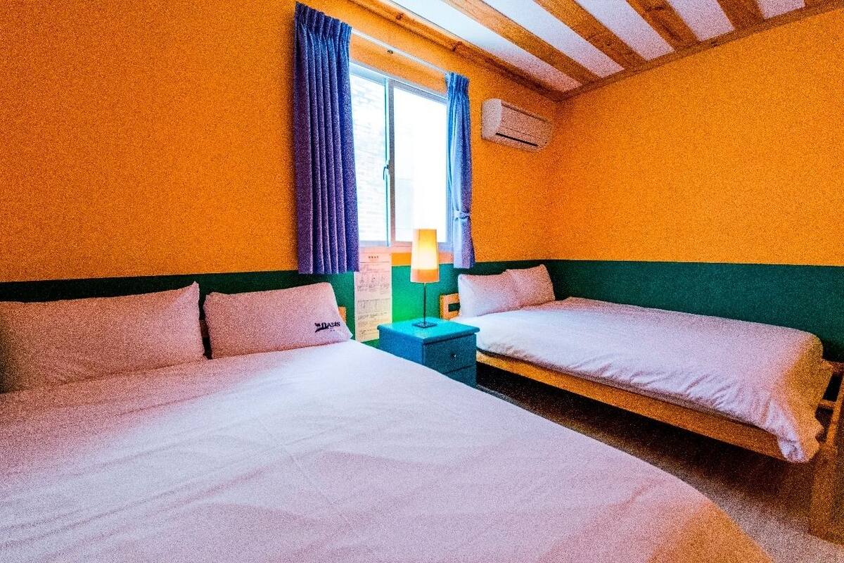 Quarto com duas camas, mesa de cabeceira, janela com cortinas e ar condicionado.