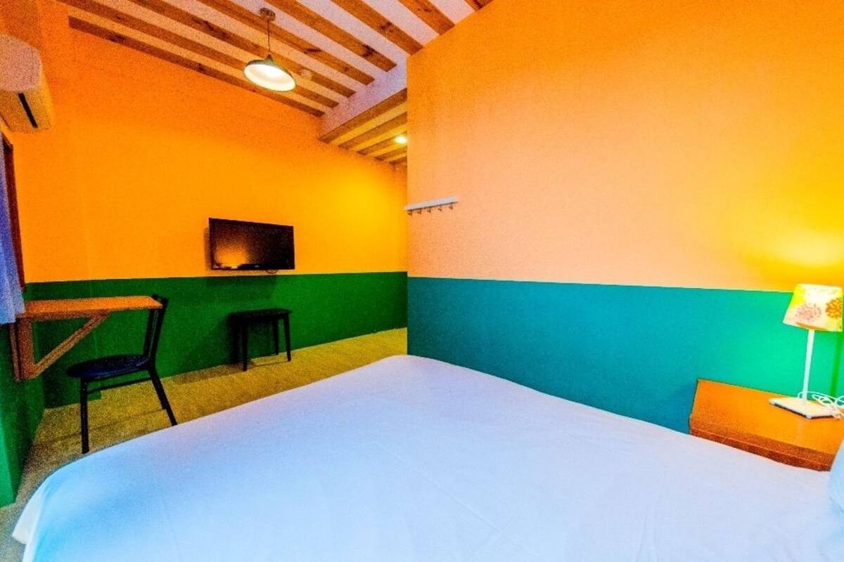 Quarto com cama, cadeira, mesinha, televisão e luminária.