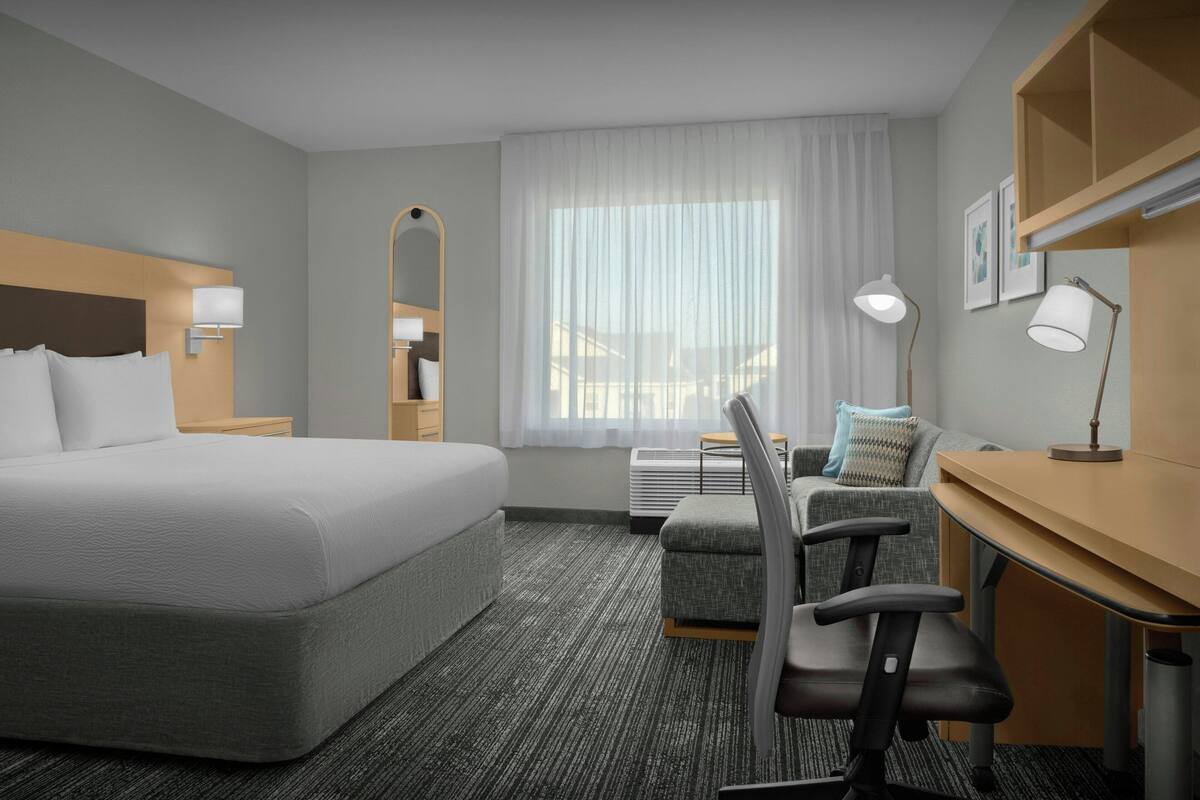 A hotel room with a large bed, a desk with a chair, a mirror, a window with curtains, and a wall-mounted lamp.