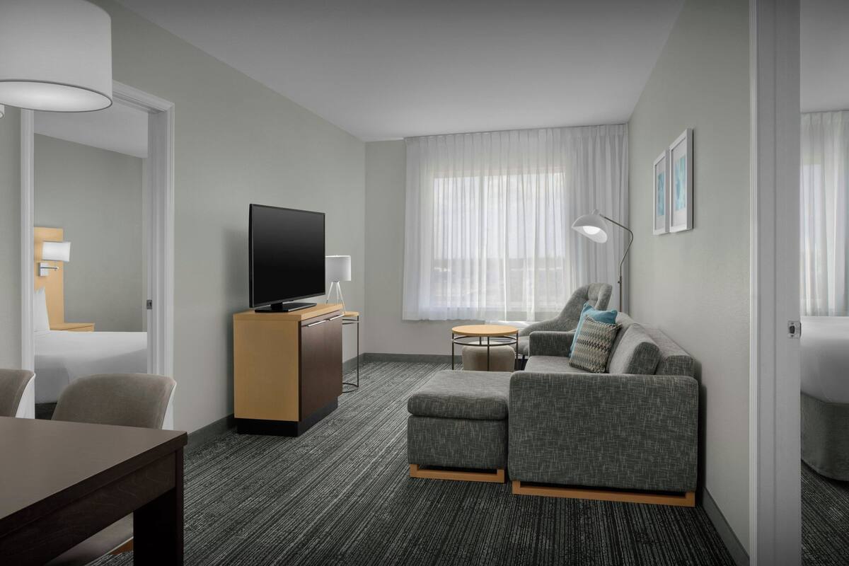 A modern hotel room with a flat-screen TV, a sofa, a small round table, and a desk with a lamp.