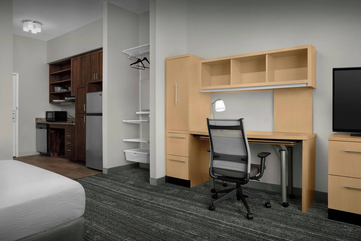 A compact hotel room with a bed, kitchenette, wooden wardrobe, desk with chair, and a TV.