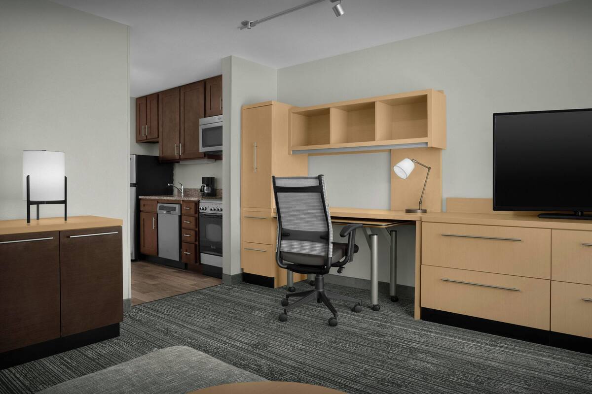 A compact hotel room with a kitchenette, a desk with a chair, a television, and a lamp.
