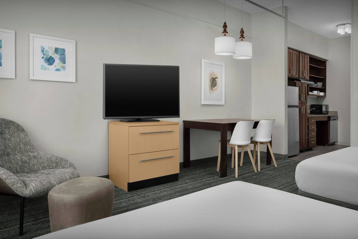 A modern hotel room with a flat-screen TV, a wooden cabinet, a dining table with chairs, and a bed.
