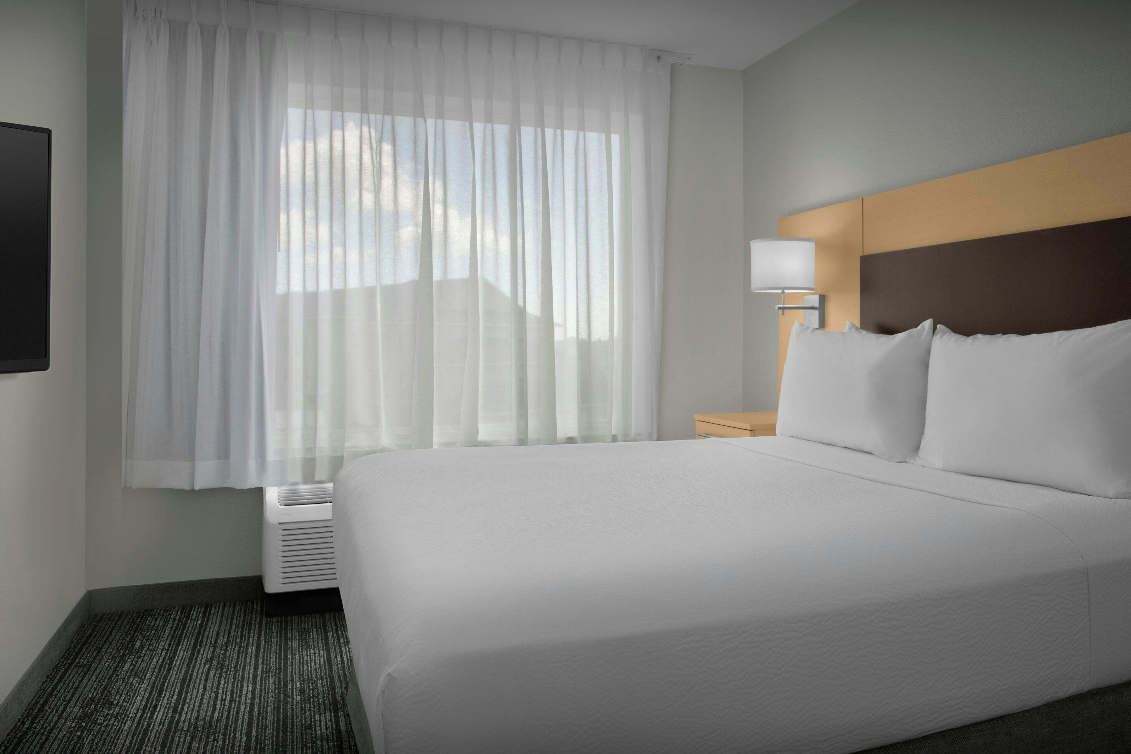 Ein Hotelzimmer mit einem großen Bett, einer Nachttischlampe und einem Fenster mit durchsichtigen Vorhängen.