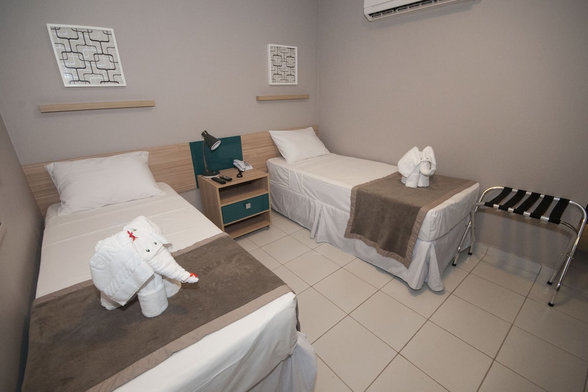 A hotel room with two beds, a nightstand, a small table, and a towel rack.