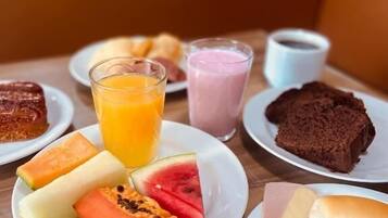 Een ontbijttafel met borden eten, waaronder fruit, een glas sinaasappelsap, een smoothie en een plakje cake.