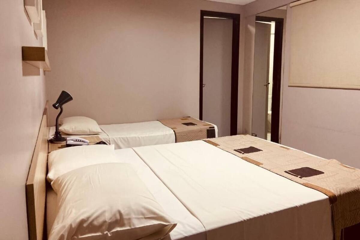 A hotel room with two beds, a bedside table with a lamp, a wardrobe, and a door leading to another room.