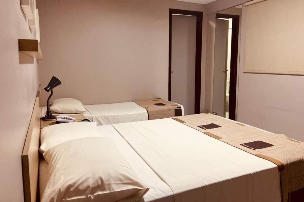 A hotel room with two beds, a bedside table with a lamp, a wardrobe, and a door leading to another room.