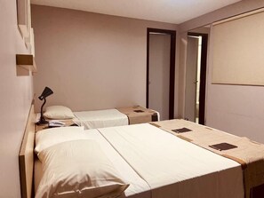 A hotel room with two beds, a bedside table with a lamp, a wardrobe, and a door leading to another room.