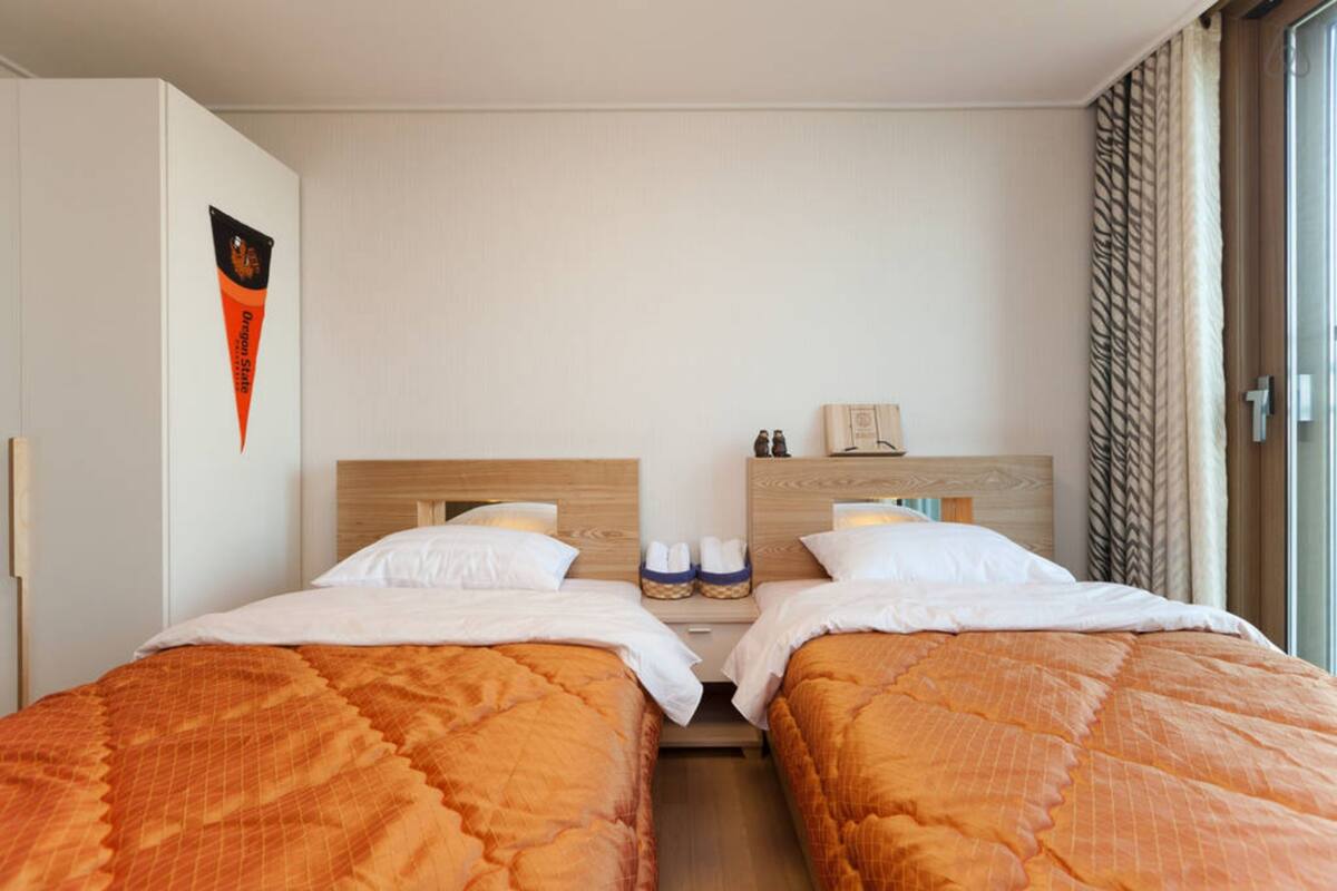 Habitació d'hotel amb dos llits, un panell de capçalera de fusta, una tauleta de nit amb una làmpada i una finestra amb cortines.
