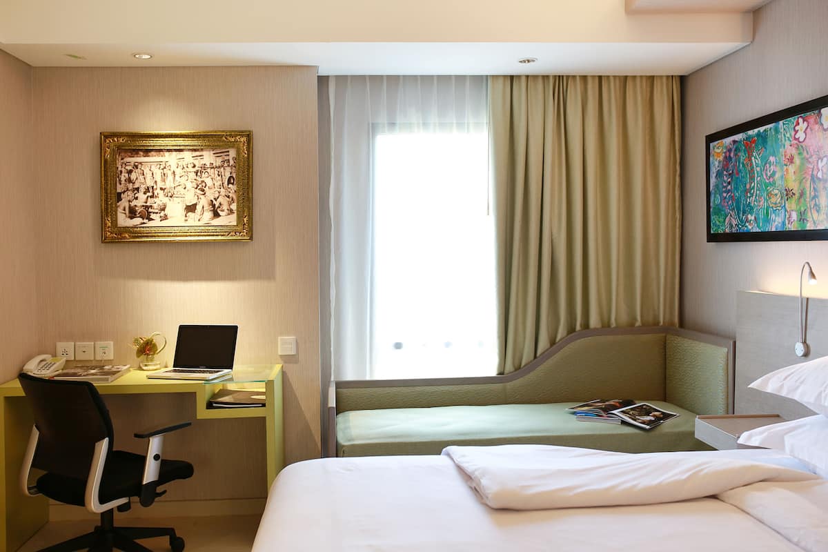 A hotel room with a desk, a bed, a window with curtains, a painting on the wall, and a lamp.