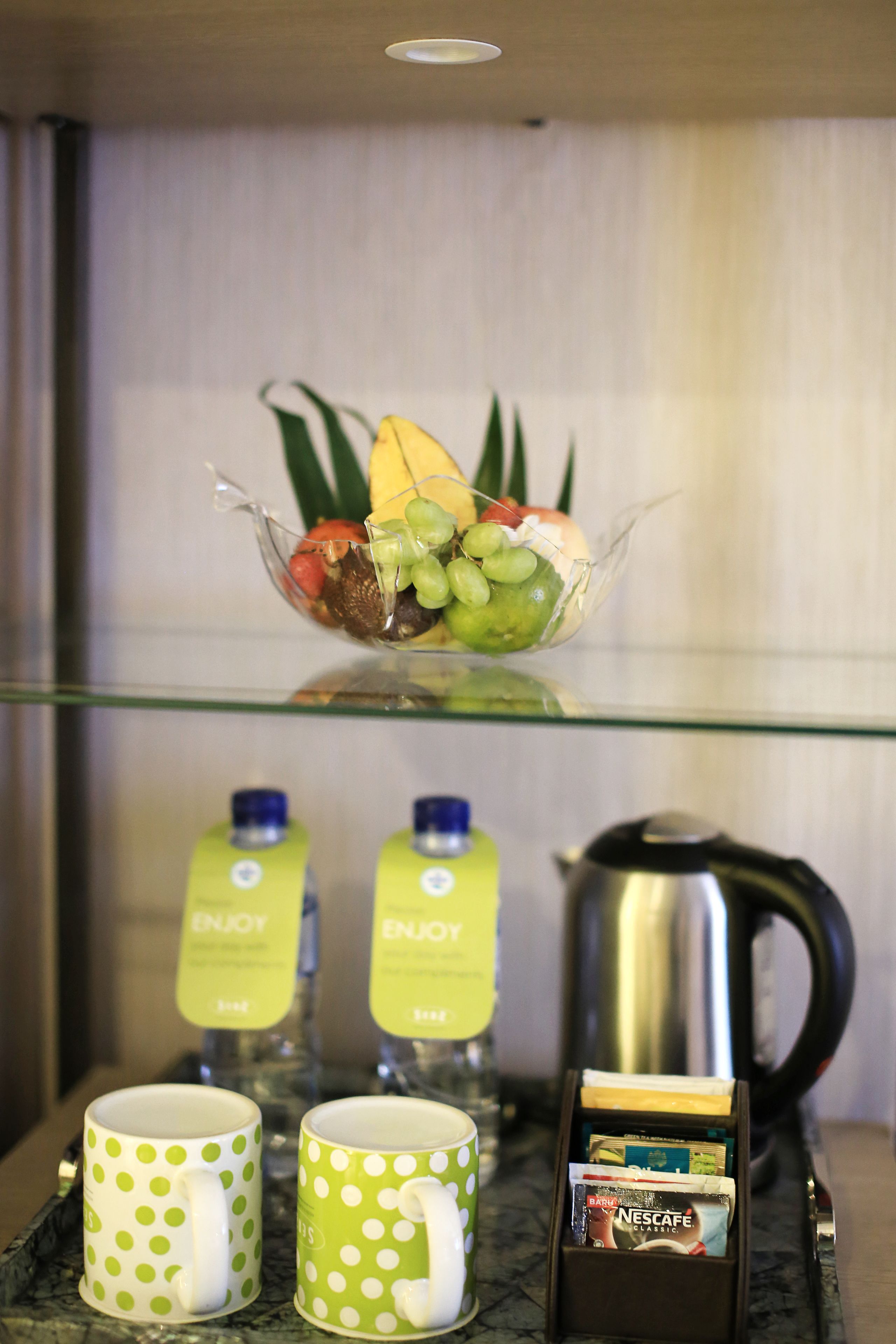 Une étagère en verre sur laquelle repose un panier de fruits contenant des raisins, une banane et d’autres fruits. En dessous, on trouve deux tasses vertes à pois, deux bouteilles d’eau et une dosette de café Nescafé.