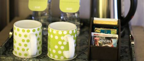 A glass shelf with a fruit bowl containing grapes, a banana, and other fruits. Below, there are two green polka-dotted mugs, two bottles of water, and a Nescafe coffee pod.