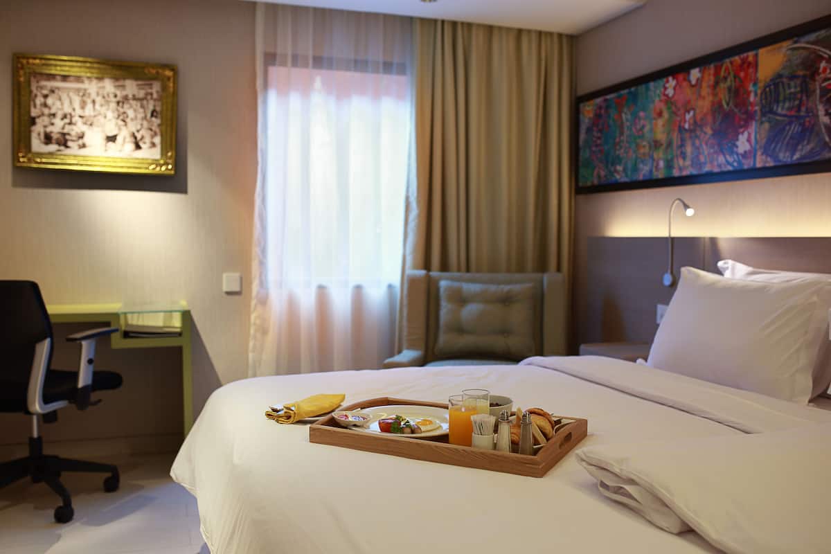 A hotel room with a neatly made bed, a desk with a chair, a tray with breakfast items, and a painting on the wall.