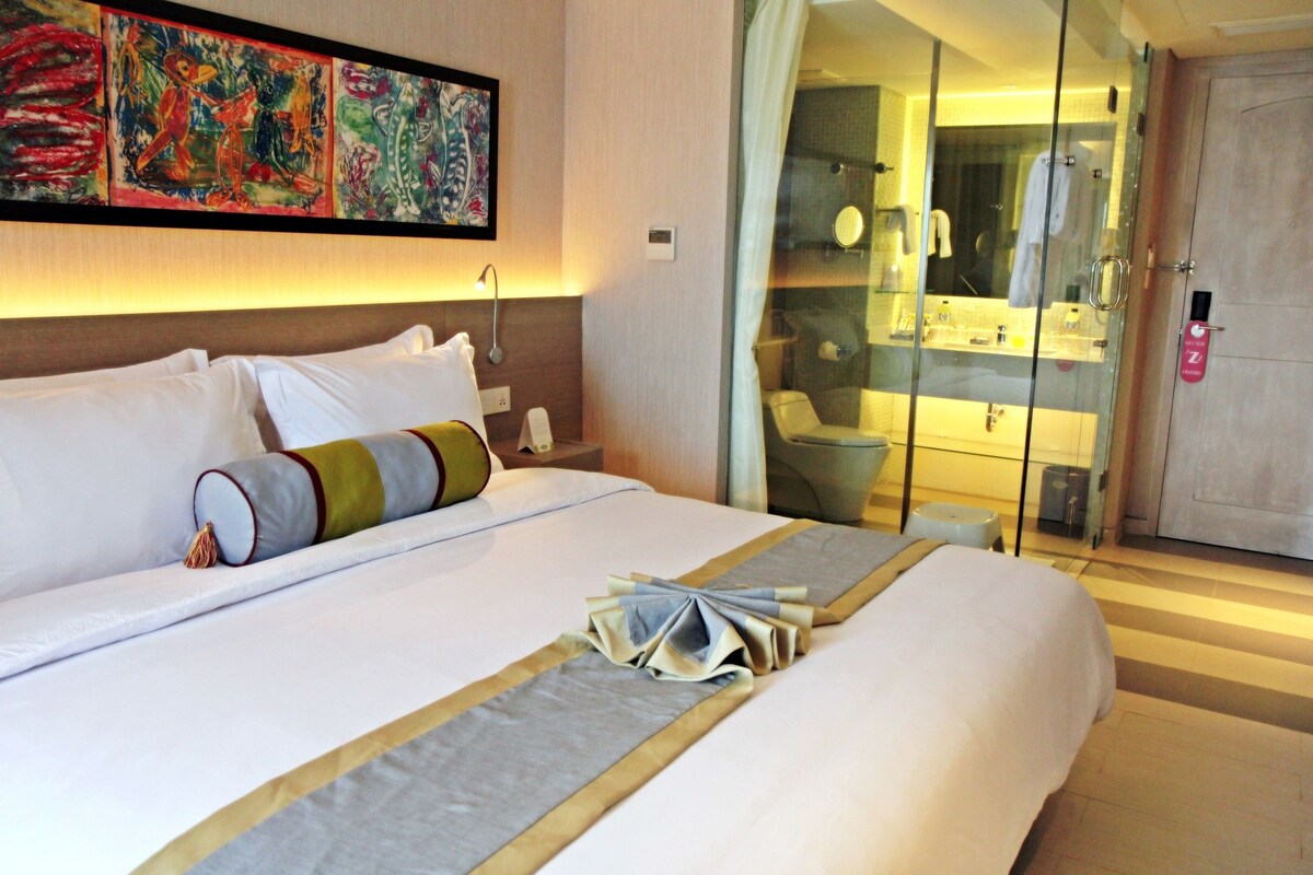A hotel room with a neatly made bed, a glass-enclosed bathroom, and a large abstract painting on the wall.