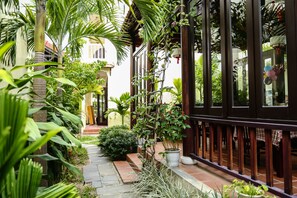 A narrow pathway flanked by lush greenery and a building with a dark wooden railing. - Hoi An Holiday Villa (Da Nang)