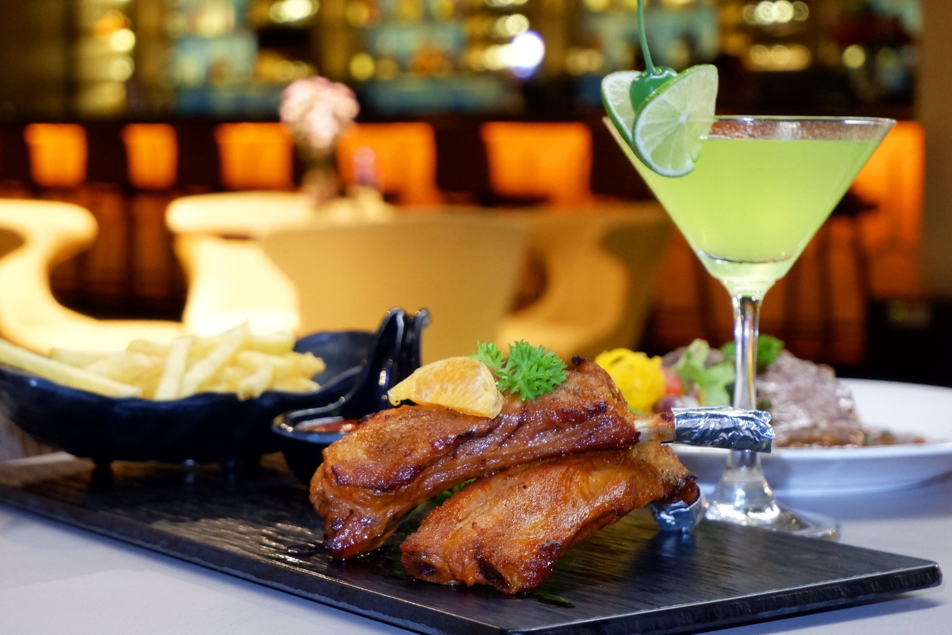 A plate of grilled meat with a garnish, a martini glass with a lime wedge, and a bowl of fries.