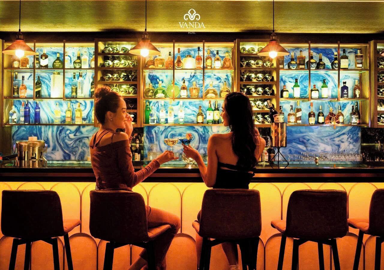 Zona de bar con un letrero del Vanda Hotel, estantes llenos de botellas de licor y dos personas sentadas en la barra.