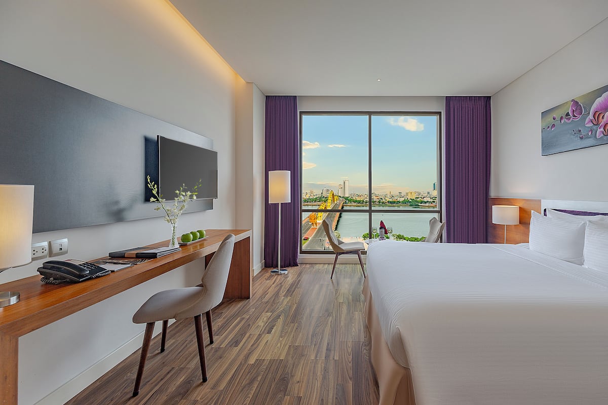 Habitación de hotel con una cama grande, un escritorio con silla, un televisor montado en la pared y vistas a la ciudad y al río.
