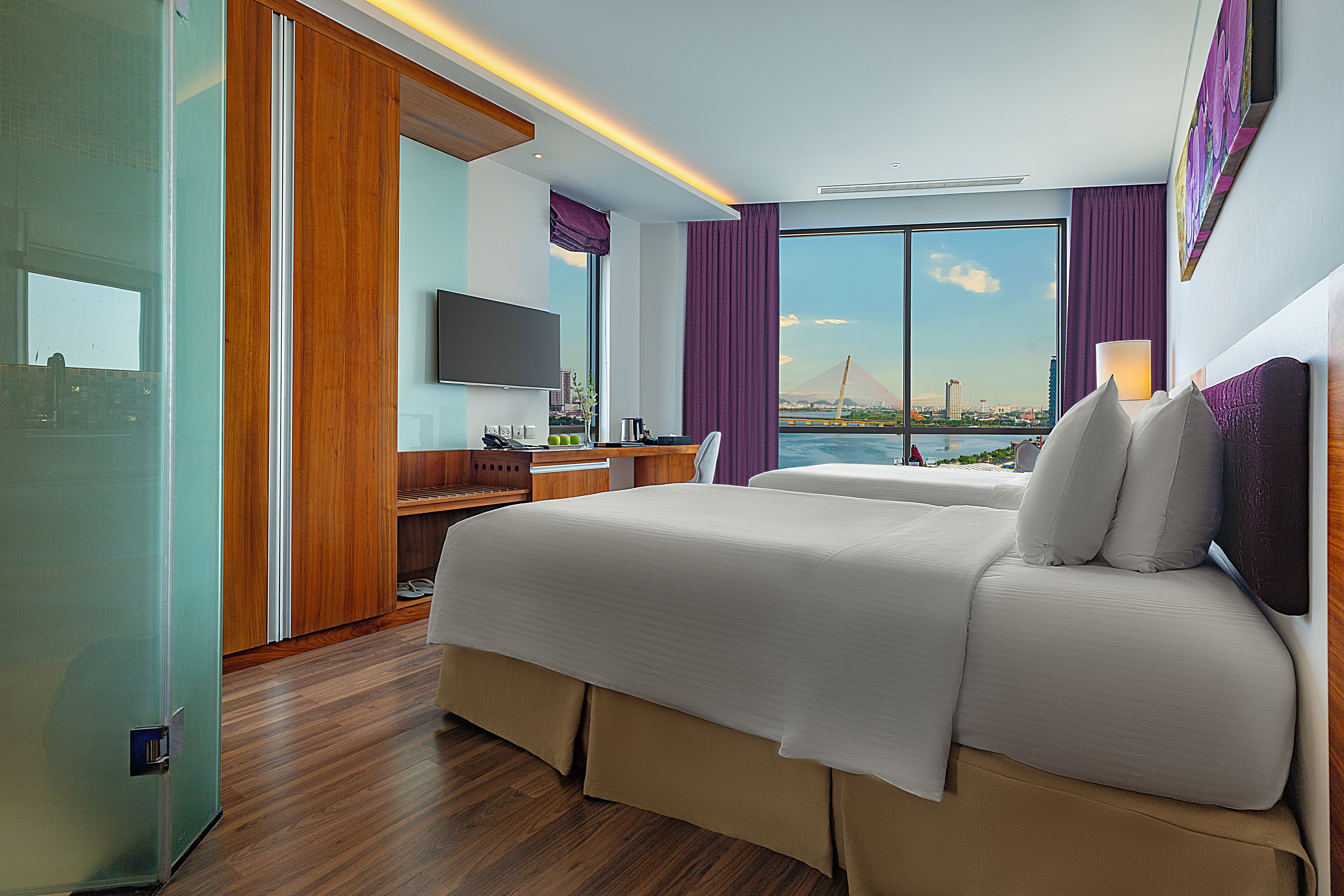 A hotel room with a large bed, a desk with a computer, a TV, and a view of a cityscape and water.