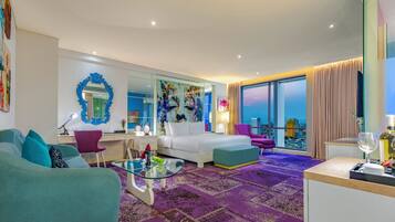 A modern hotel room with a large bed, a sofa, a glass coffee table, and a view of the city.