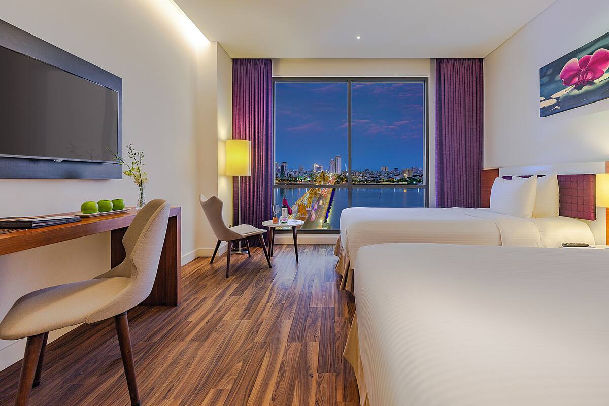 Habitación de hotel con una cama grande, un escritorio con silla, un televisor y una ventana con vista a la ciudad.