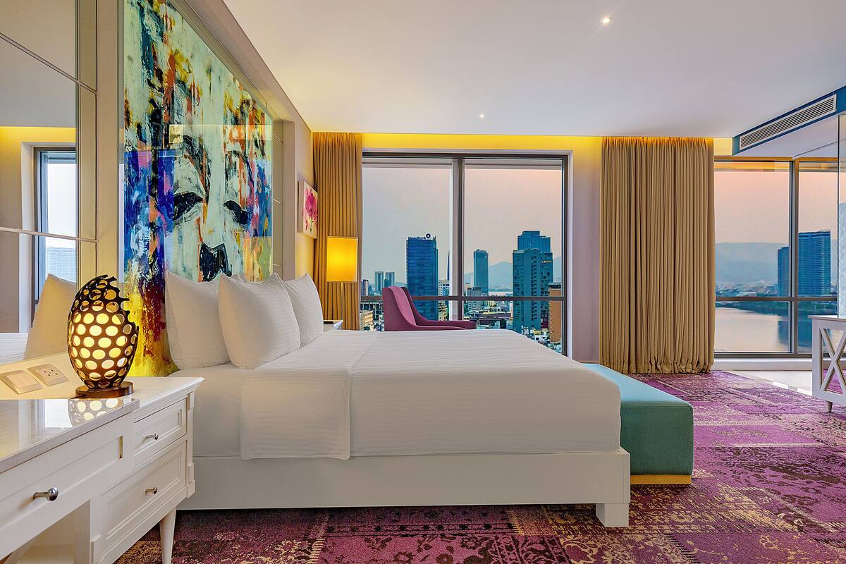 Habitación de hotel con una cama grande, un escritorio con lámpara, una silla y vistas a la ciudad.