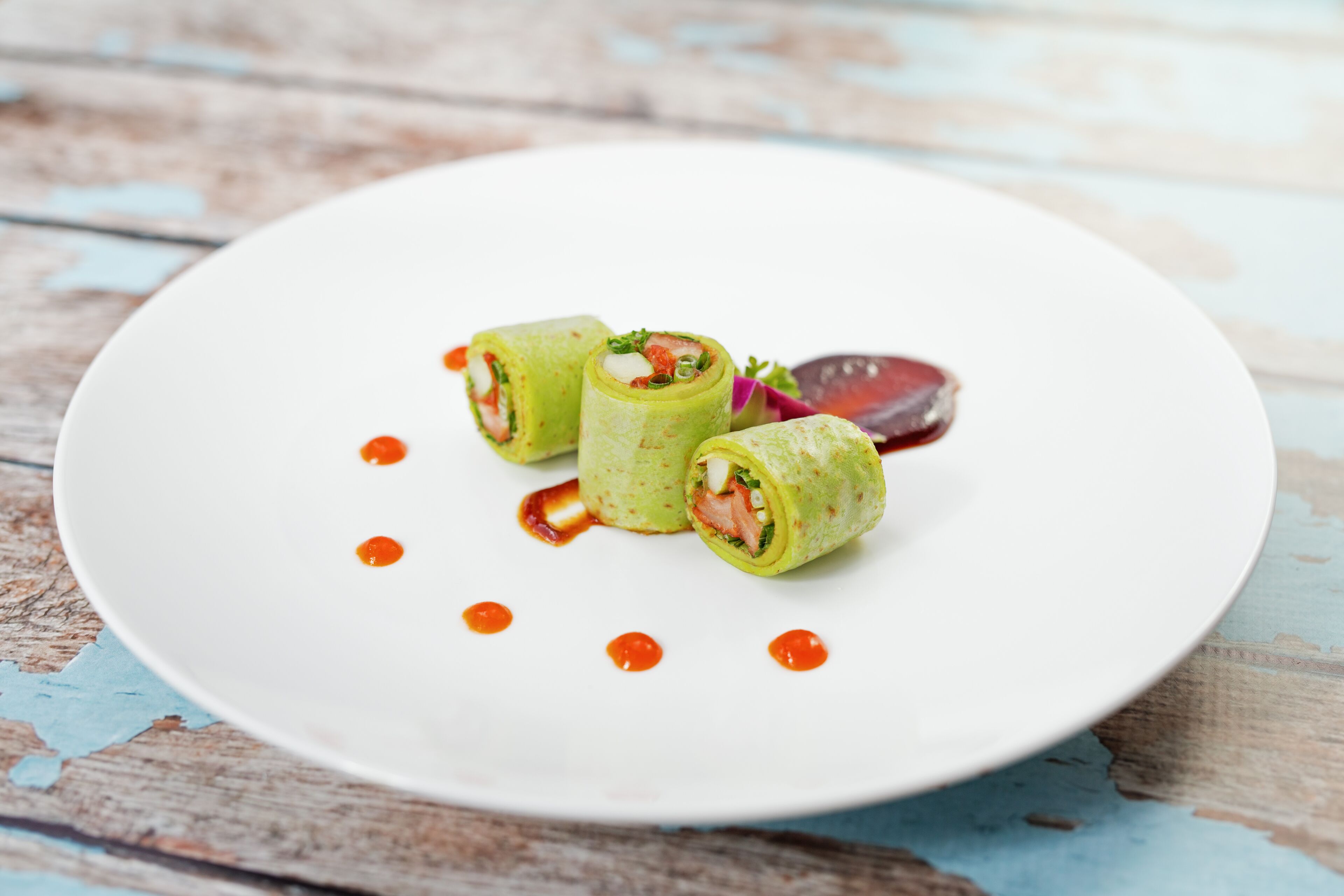 Une assiette blanche sur laquelle sont disposés trois amuse-gueules roulés et une tranche de betterave, le tout posé sur une table en bois rustique.