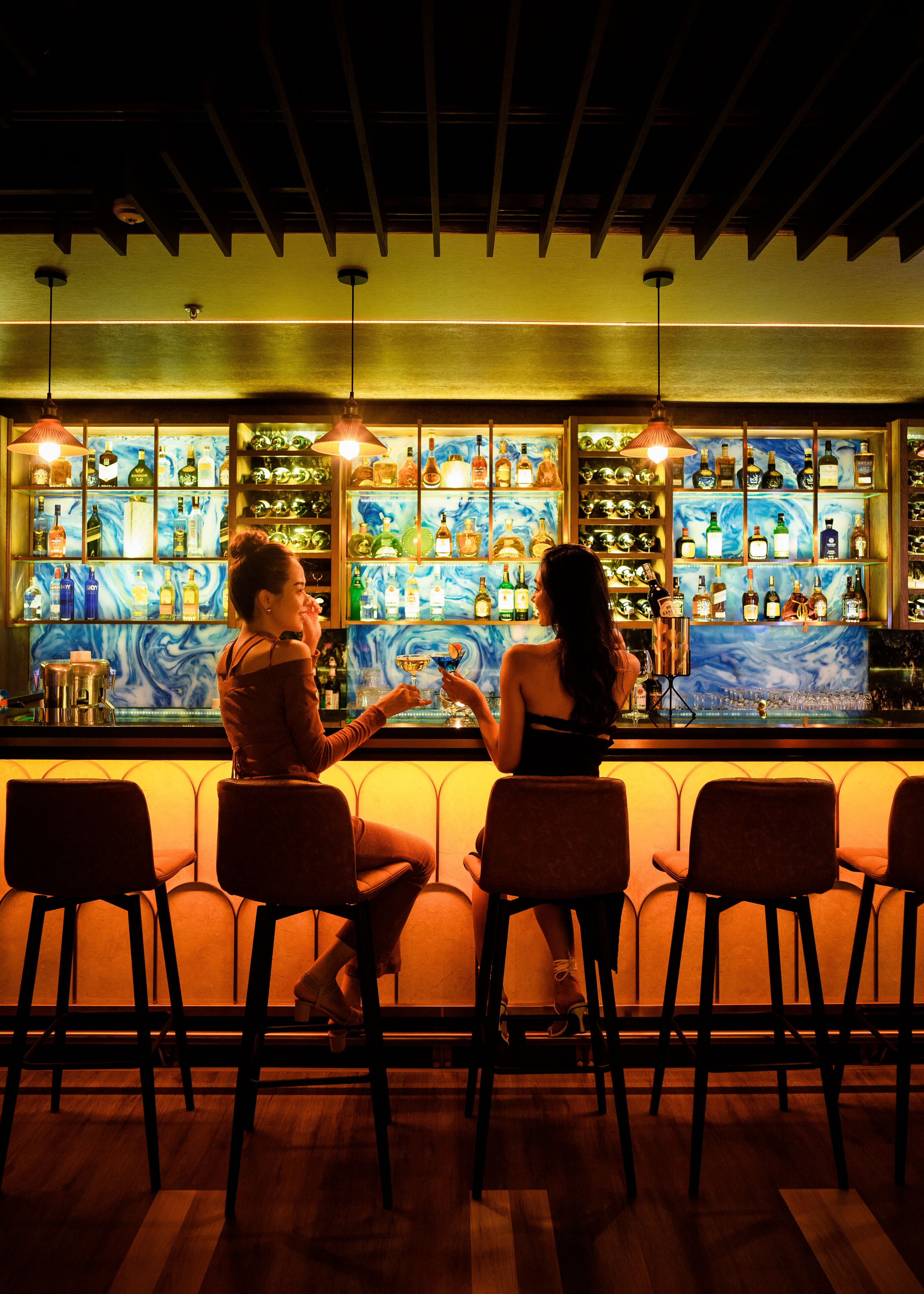 A bar with two patrons seated at the bar, a wooden ceiling, and shelves stocked with bottles.