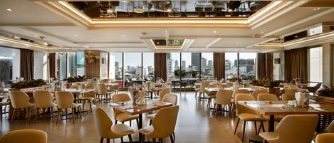 Un'ampia sala da pranzo con diversi tavoli e sedie, grandi finestre che offrono vista sulla città e un moderno design del soffitto.